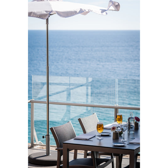 Outdoor dining table with chairs and drinks on a balcony with a Pamela parasol overlooking the ocean.